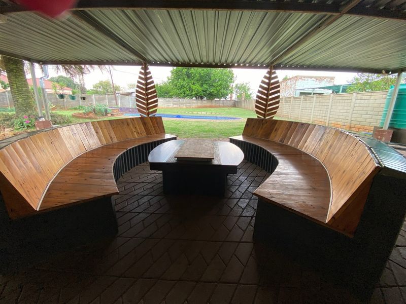 A covered outdoor patio area featuring built-in curved wooden seating, overlooking a private garden and swimming pool.