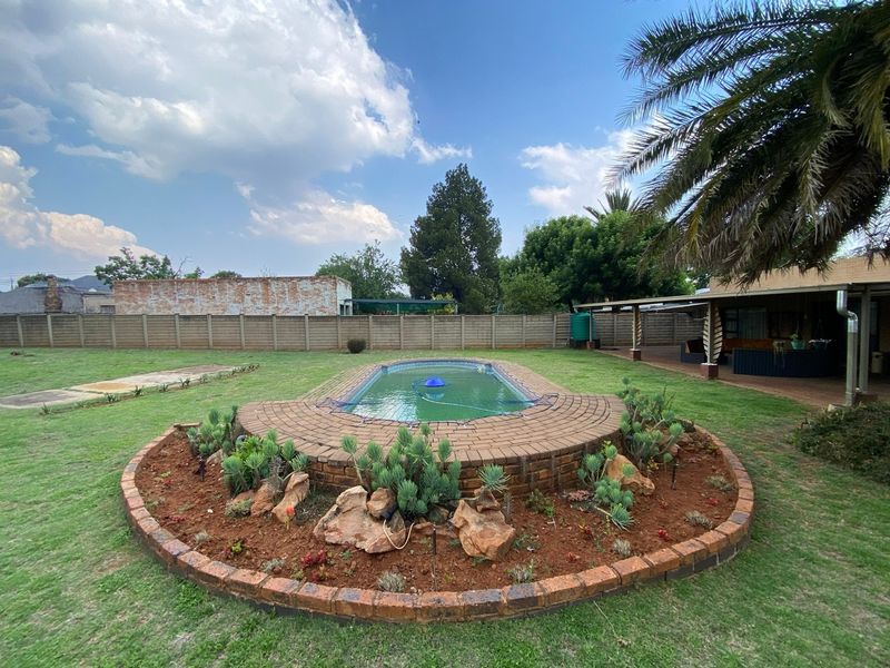Spacious backyard featuring a central swimming pool with a brick surround, landscaped garden beds, and a covered patio area.