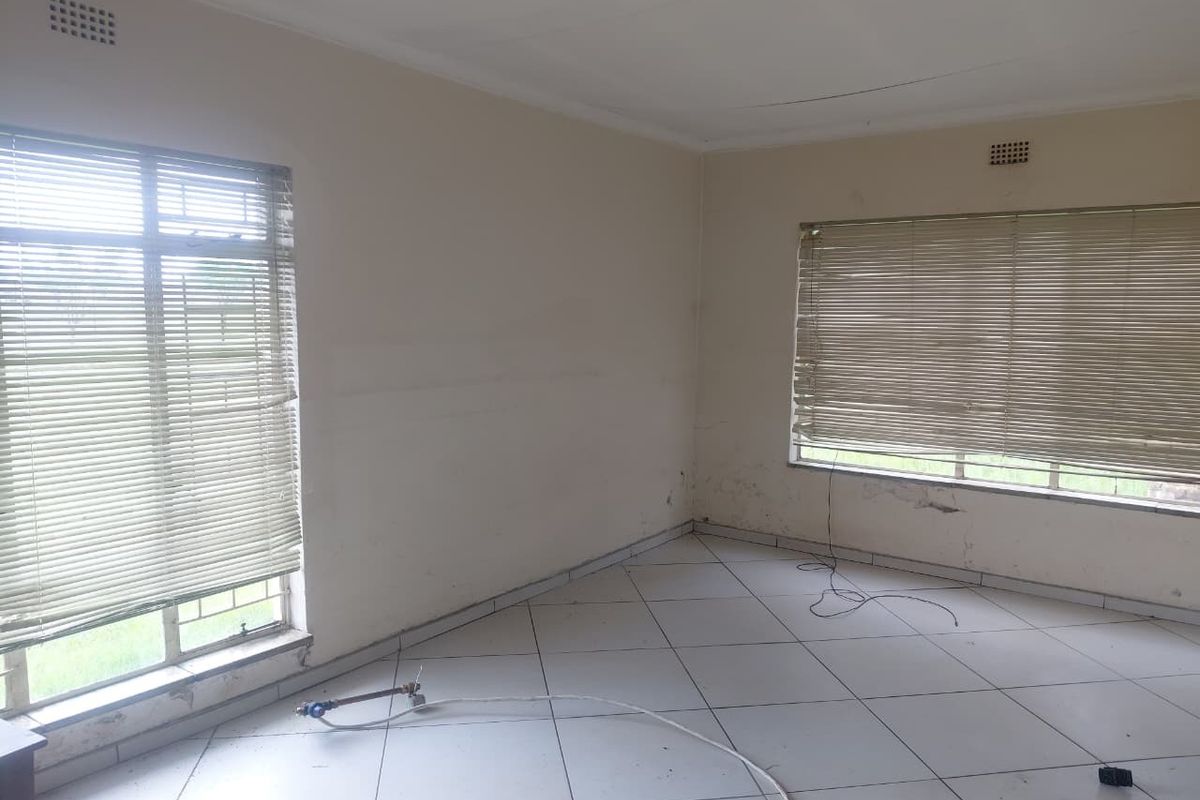 An empty living room featuring tiled flooring, ceiling fan, and venetian blinds, showing signs of wall plaster damage and damp staining.