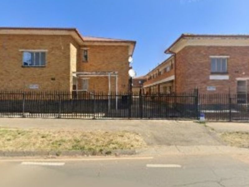 Street view of a multi-unit apartment complex featuring brick facades, tiled roofing, and secure metal perimeter fencing.