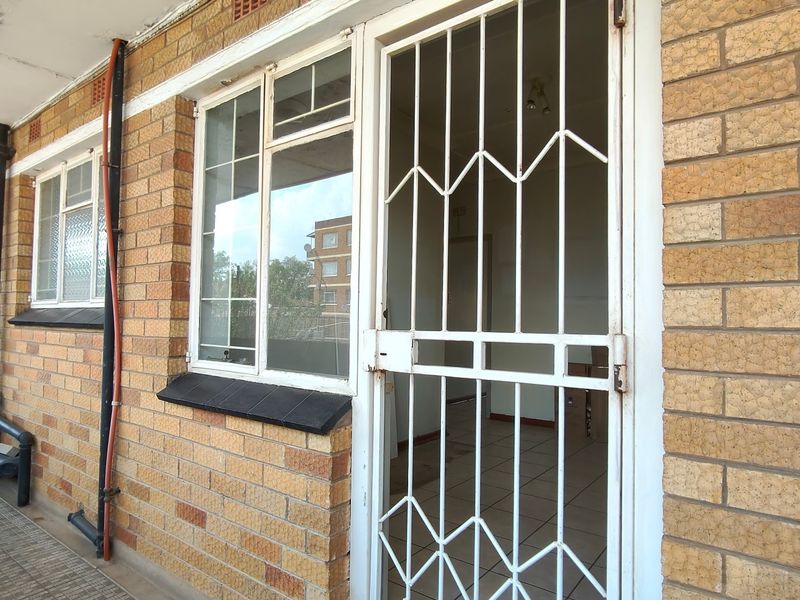 Exterior view of an apartment entrance featuring a brick facade, security gate, and window, showing signs of wear and peeling paint on the ceiling.
