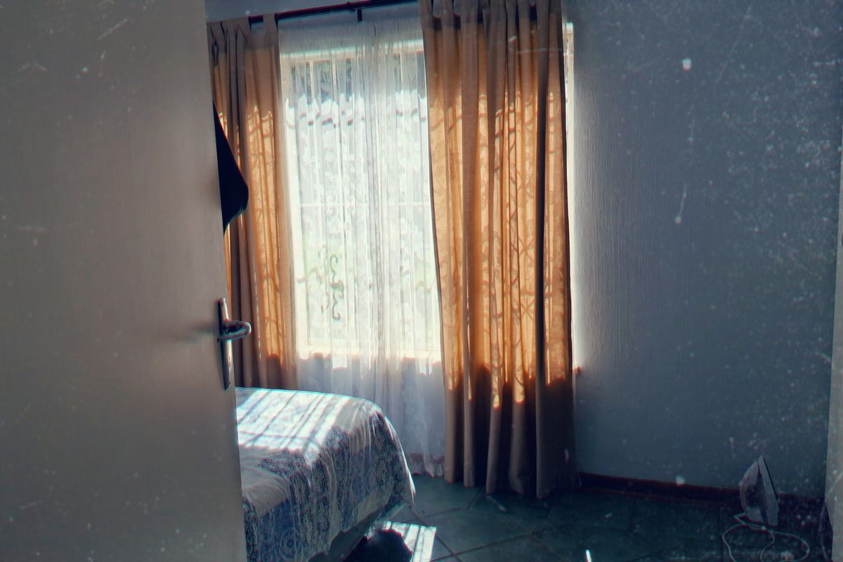 A bedroom featuring tiled flooring and a built-in cupboard, illuminated by natural light through a window with curtains.