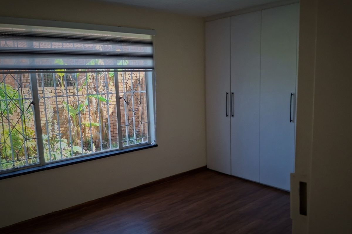 A bright, empty bedroom featuring modern laminate flooring, built-in cupboards, and large windows fitted with security bars and roller blinds.