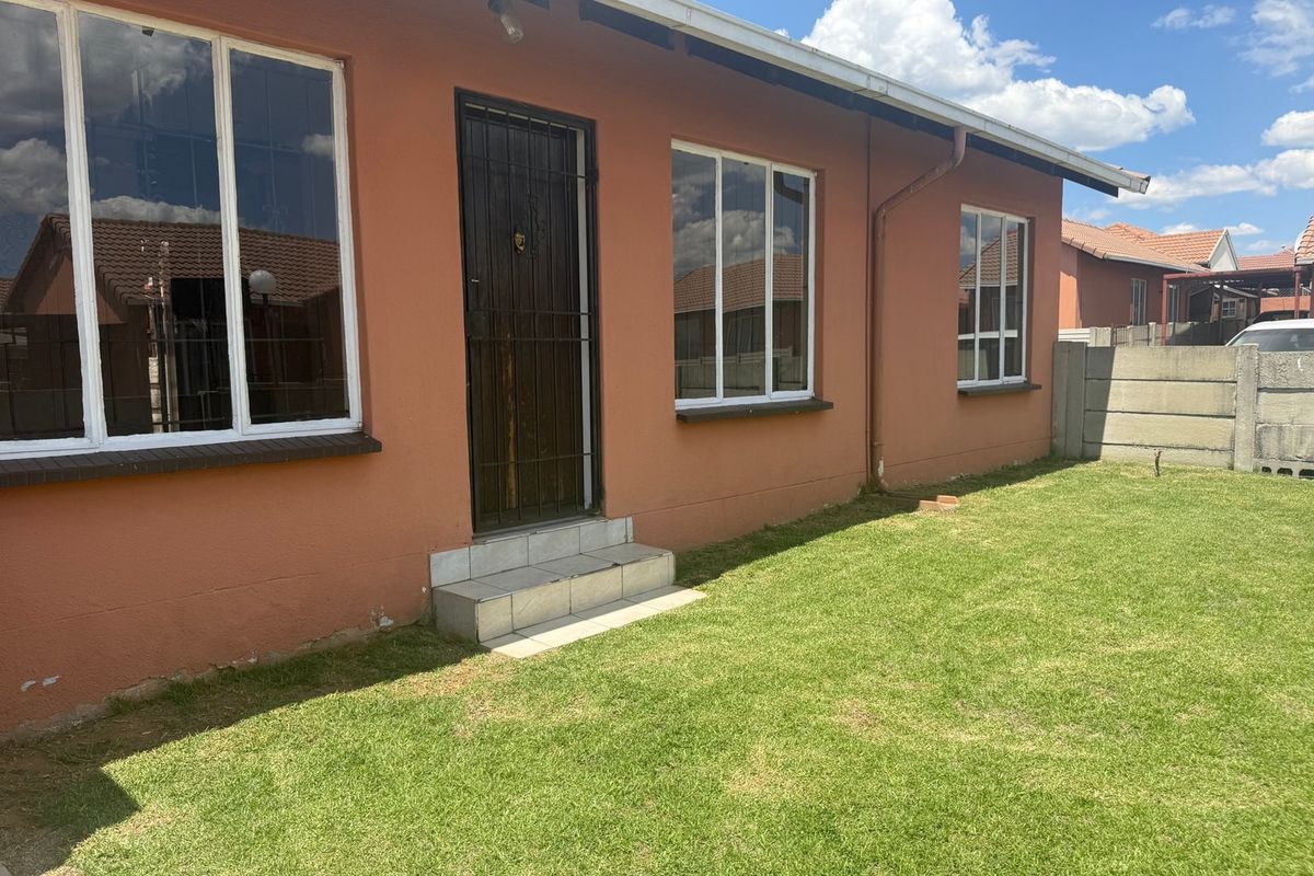 Rear exterior view of the house featuring a small tiled entrance, aluminium windows, and a private lawn area enclosed by a precast concrete wall.