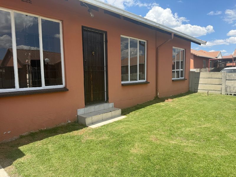 Rear exterior view of the house featuring a small tiled entrance, aluminium windows, and a private lawn area enclosed by a precast concrete wall.
