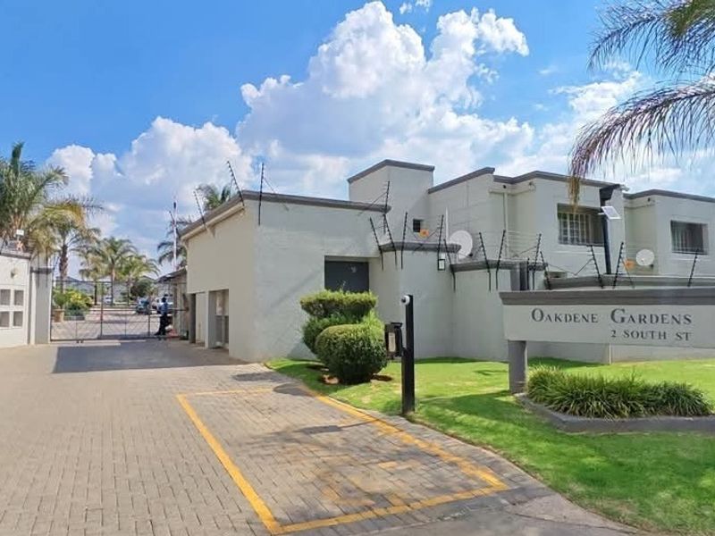 Front entrance view of Oakdene Gardens townhouse complex featuring a paved driveway, secure gated access, and well-maintained perimeter electric fencing.