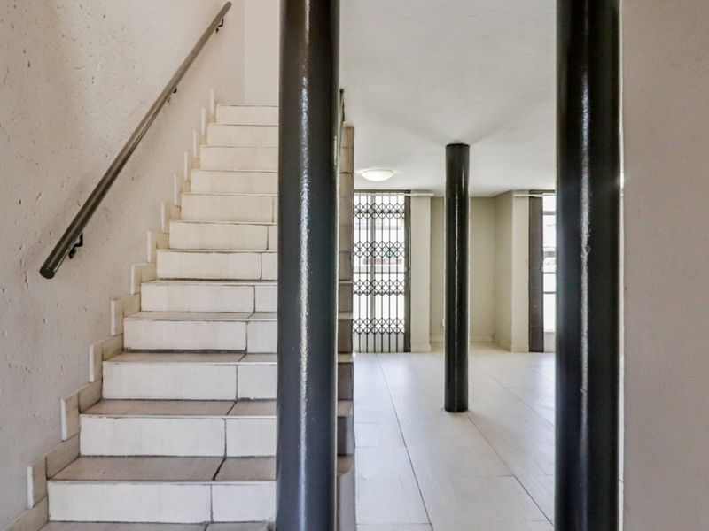 An open-plan hallway featuring a tiled staircase with a metal handrail, structural support columns, and a security gate leading to the exterior.
