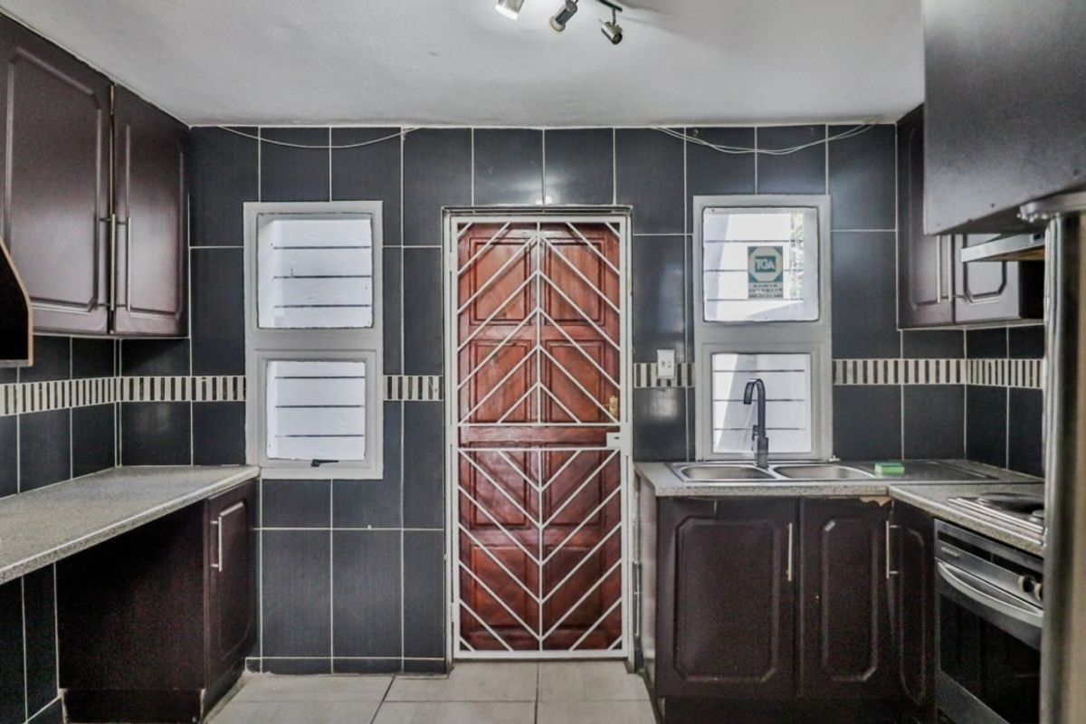Functional kitchen featuring dark wood cabinetry, stone-look countertops, double sink, built-in oven, and tiled splashback with direct access to an exterior security gate.
