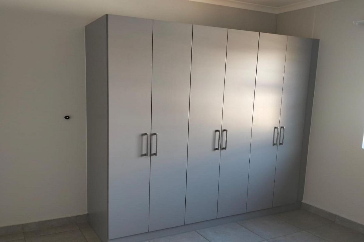 A clean, empty bedroom featuring modern grey built-in wardrobes, tiled flooring, and a window fitted with security bars.