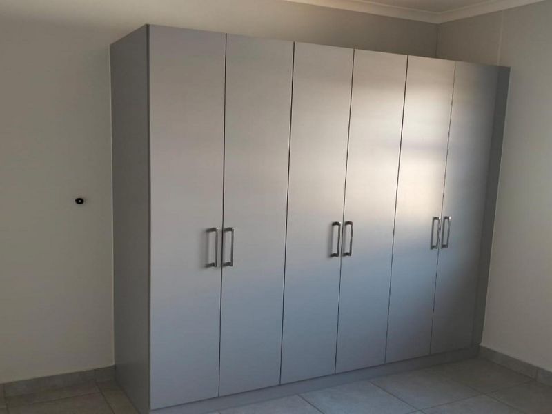 A clean, empty bedroom featuring modern grey built-in wardrobes, tiled flooring, and a window fitted with security bars.