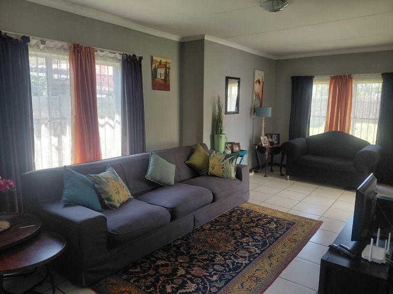 A spacious living room featuring tiled flooring, neutral wall tones, and large windows fitted with security bars, providing ample natural light.