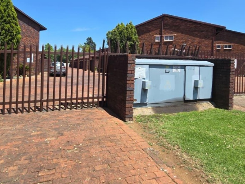 Front entrance view of the townhouse complex featuring brick paving, secure metal fencing, and a prominent electrical utility box.