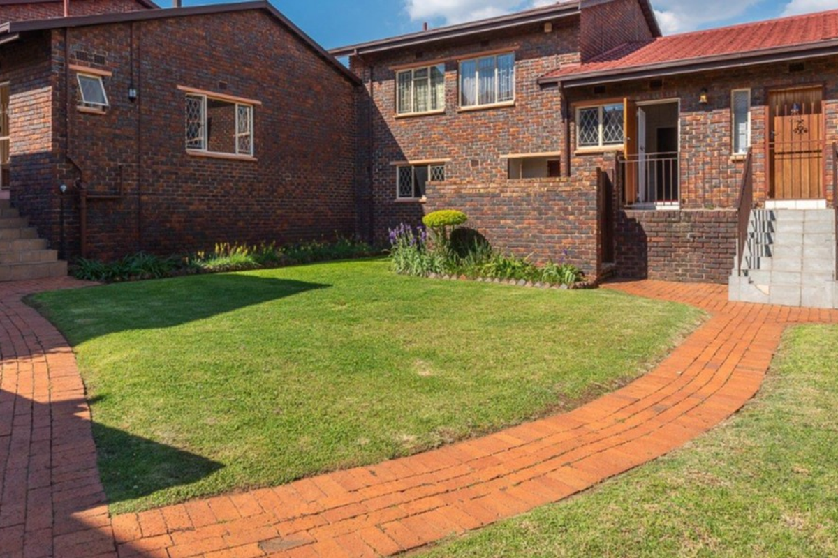 A well-maintained rear exterior view of a face brick townhouse complex featuring paved walkways, manicured lawns, and secure access points.