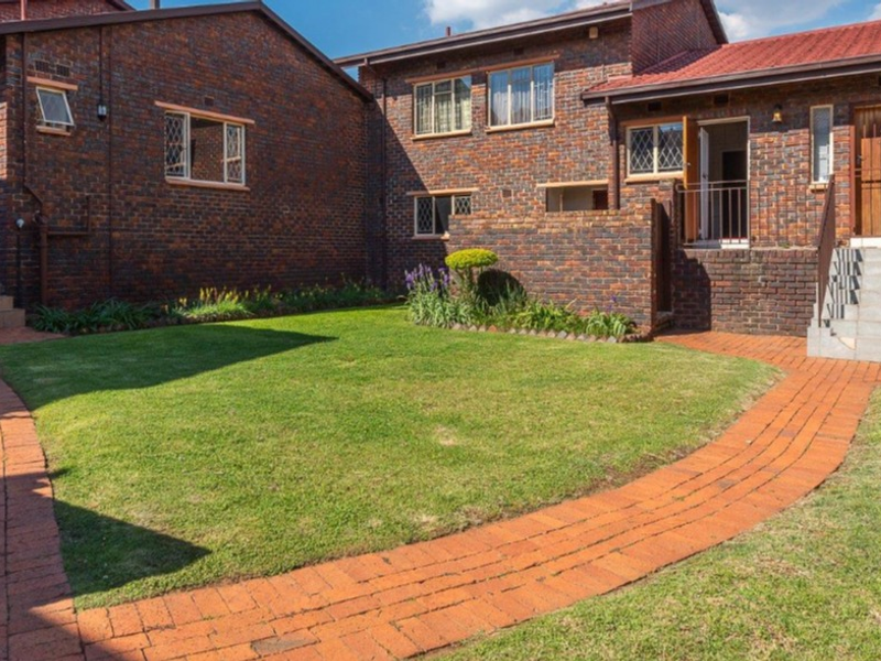 A well-maintained rear exterior view of a face brick townhouse complex featuring paved walkways, manicured lawns, and secure access points.
