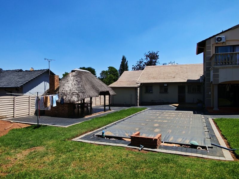 Rear exterior view featuring a swimming pool with cover, a thatched lapa entertainment area, paved patio, and a double-story house with a balcony.