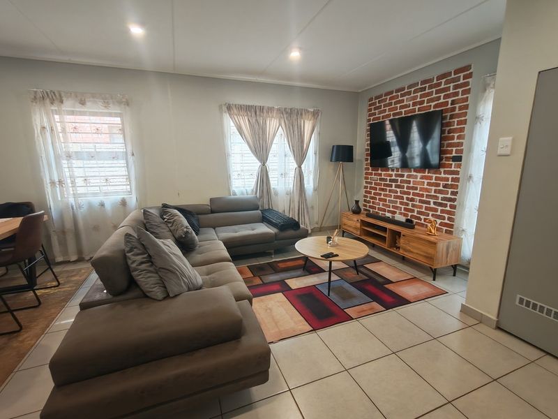 A well-maintained living room featuring a distinctive exposed brick accent wall, neutral tiled flooring, and recessed ceiling lighting, creating a warm and inviting atmosphere.