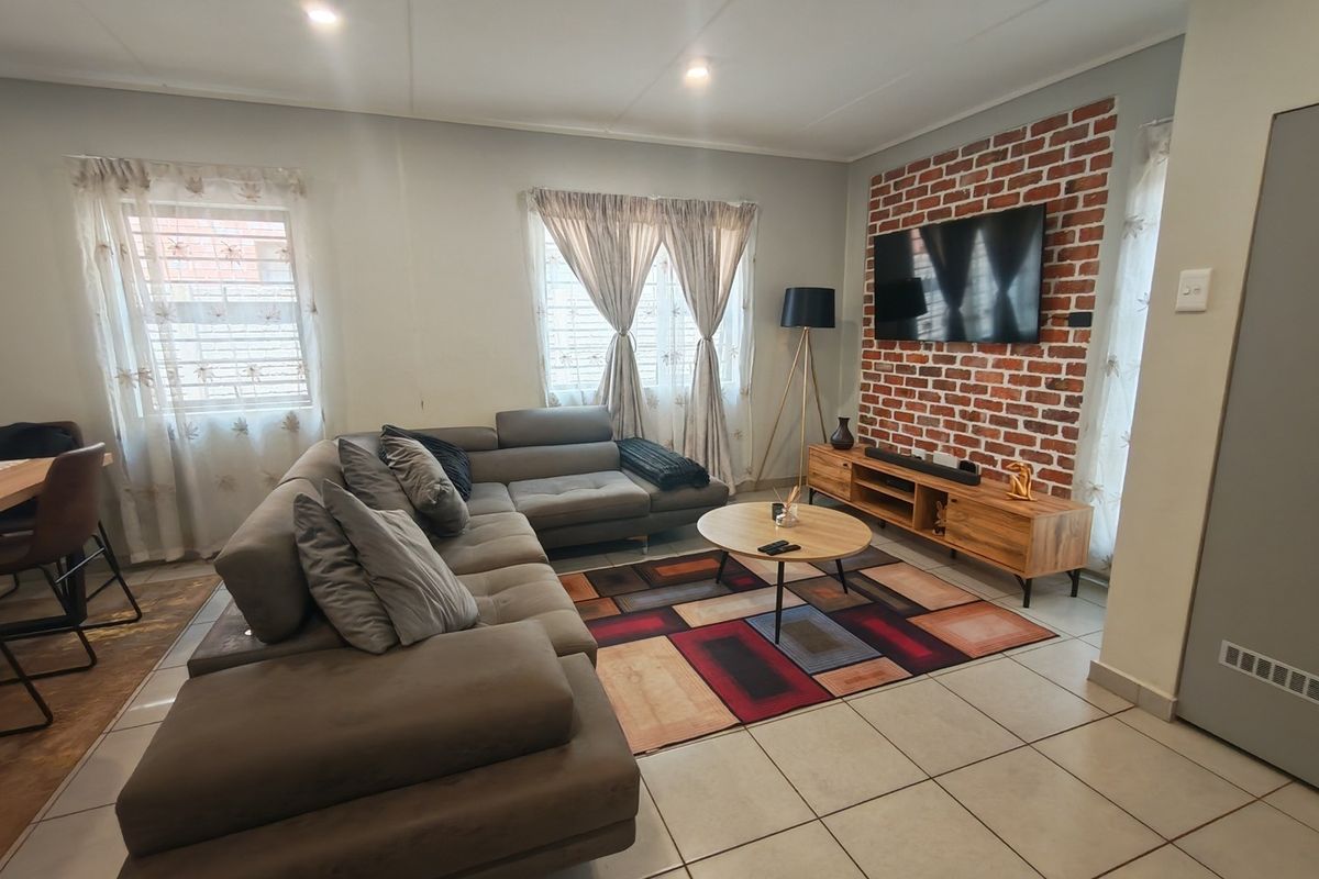 A well-maintained living room featuring a distinctive exposed brick accent wall, neutral tiled flooring, and recessed ceiling lighting, creating a warm and inviting atmosphere.