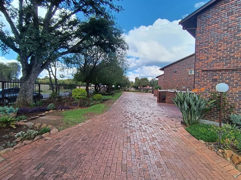 A well-maintained brick driveway leading to a face brick townhouse facade, featuring established garden beds and secure perimeter fencing.