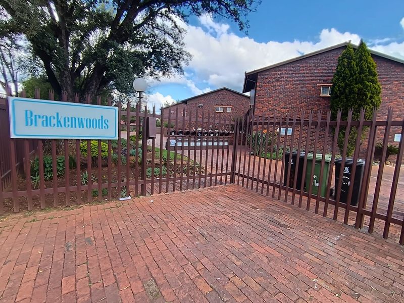 Front entrance view of the Brackenwoods townhouse complex featuring brick facade buildings, paved driveway, and secure metal perimeter fencing.