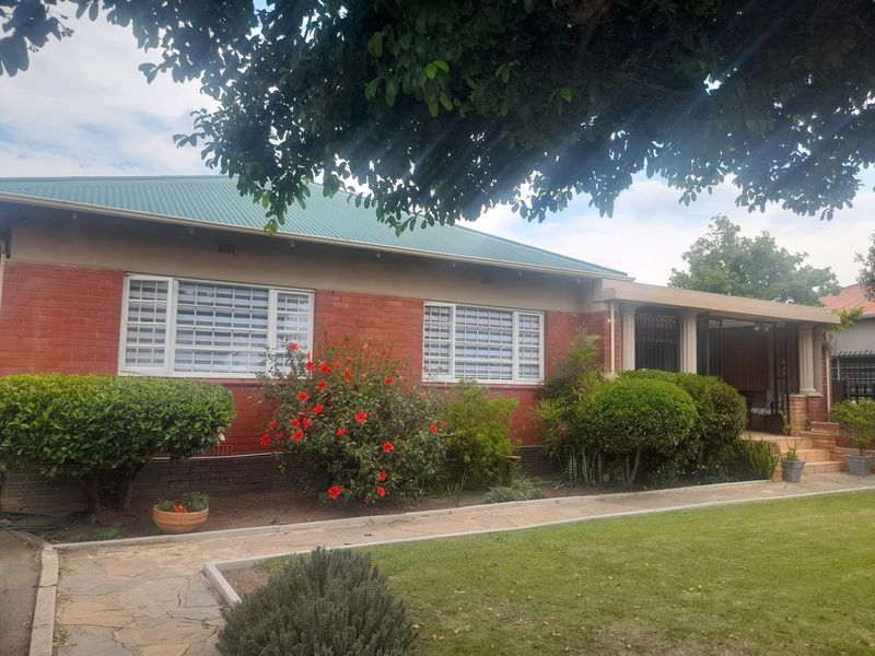Front elevation of a single-story brick house featuring a metal roof, security-barred windows, and a covered entrance porch set within a landscaped garden.