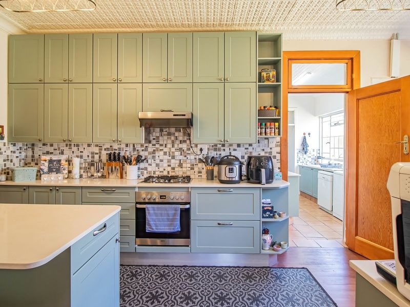 A well-maintained kitchen featuring sage green cabinetry, stone countertops, a mosaic tile backsplash, and a distinctive pressed metal ceiling, with an adjacent scullery area.