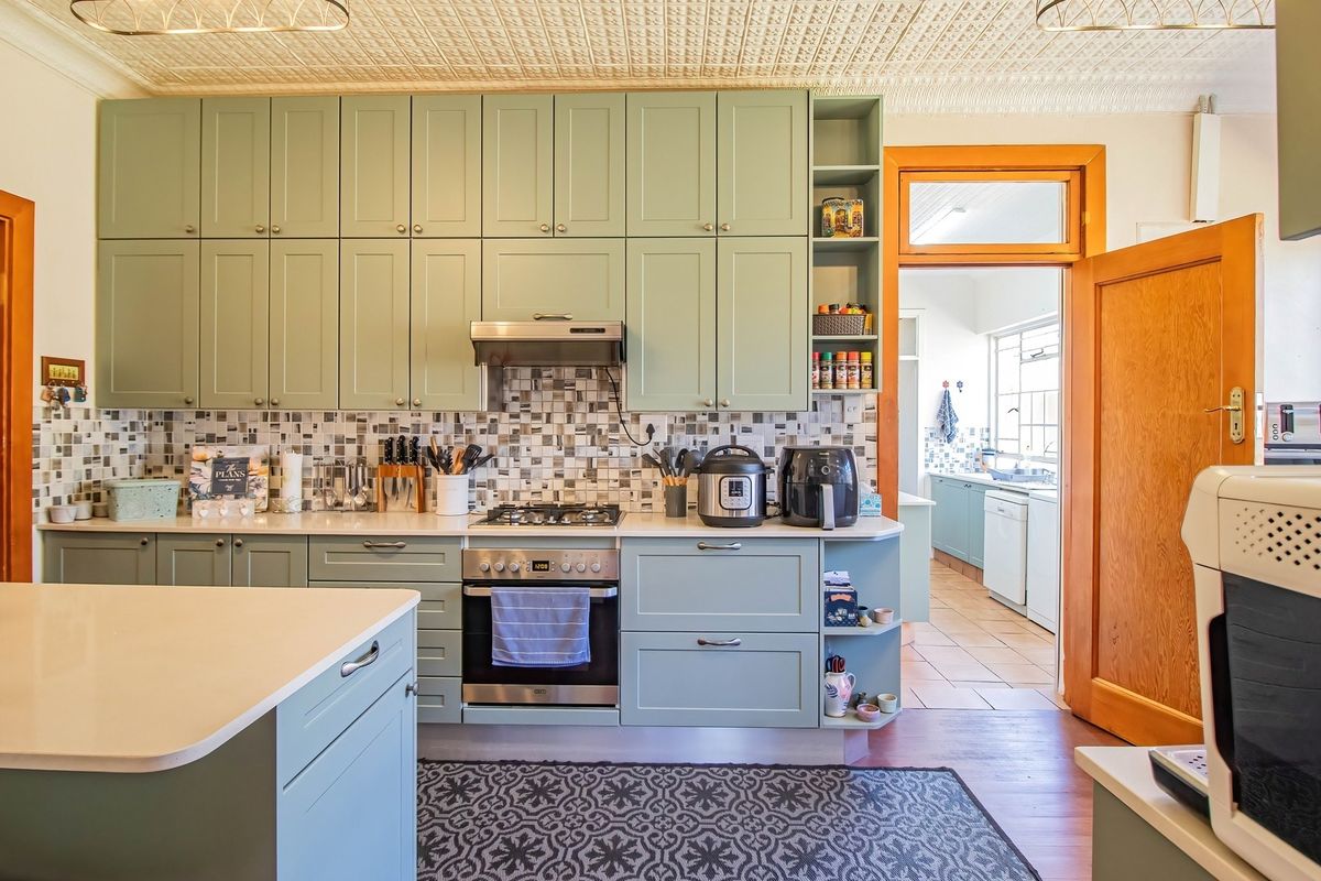 A well-maintained kitchen featuring sage green cabinetry, stone countertops, a mosaic tile backsplash, and a distinctive pressed metal ceiling, with an adjacent scullery area.