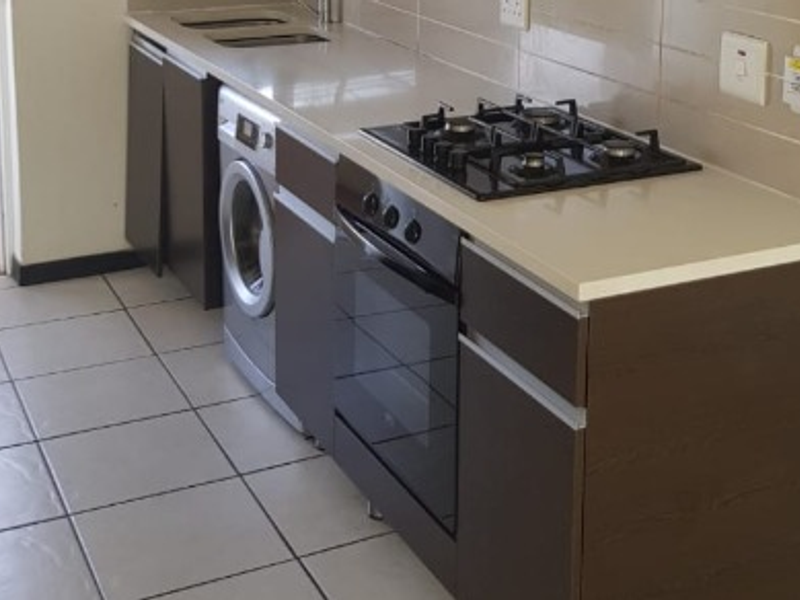 A compact, modern kitchen featuring dark cabinetry, stone countertops, a built-in gas hob, oven, and tiled flooring with direct access to an outdoor area.