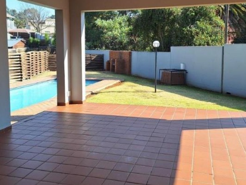 A spacious covered patio featuring tiled flooring, overlooking a private swimming pool and a well-maintained garden with a built-in brick braai area.