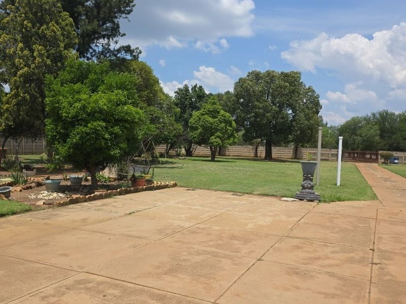 A spacious garden area featuring a large lawn, mature trees, and a paved driveway enclosed by a precast concrete boundary wall.