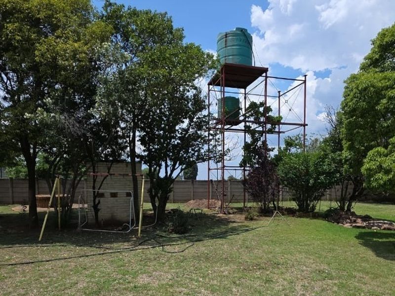 Spacious garden area featuring a raised steel water tank stand, established trees, and a brick braai structure enclosed by a precast concrete boundary wall.