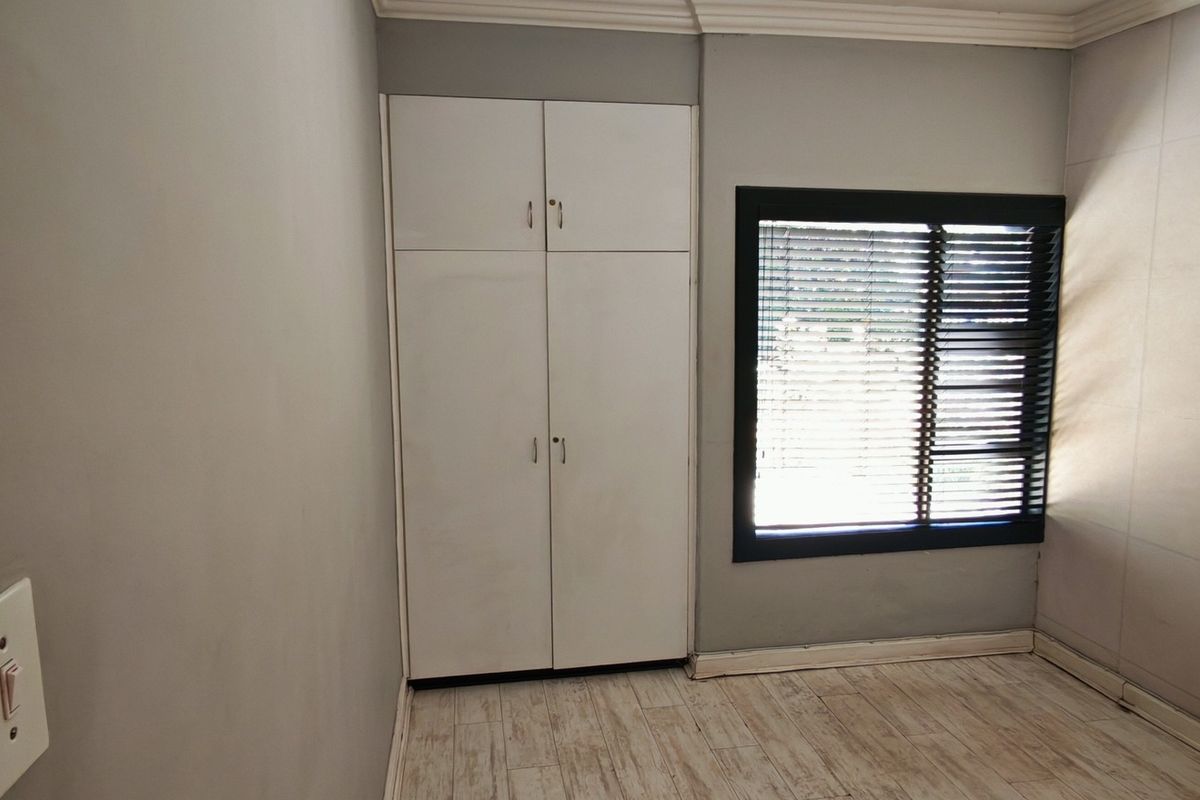A compact bedroom featuring neutral tiled flooring, built-in cupboards, and a window with fitted blinds, offering a clean and functional space.