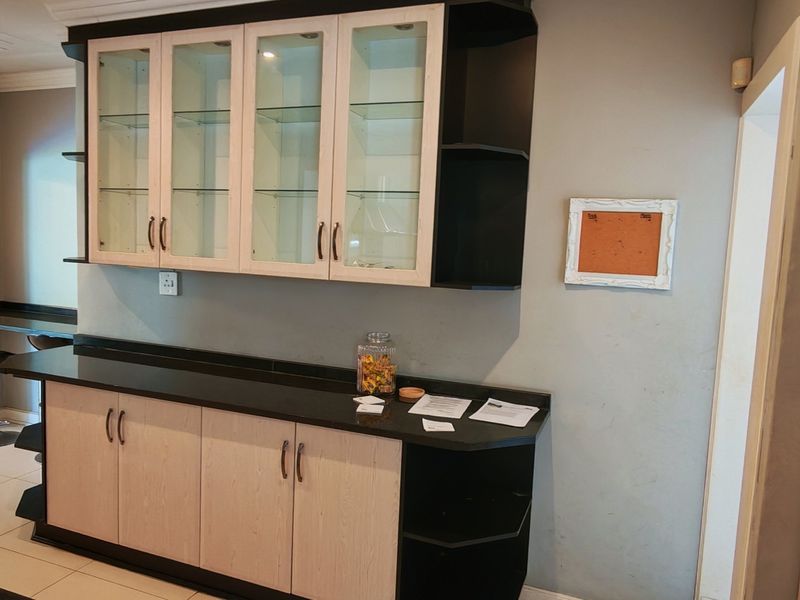A kitchen area featuring light wood-toned cabinetry with black accents, glass-fronted display units, and a breakfast bar, set against a neutral wall finish.