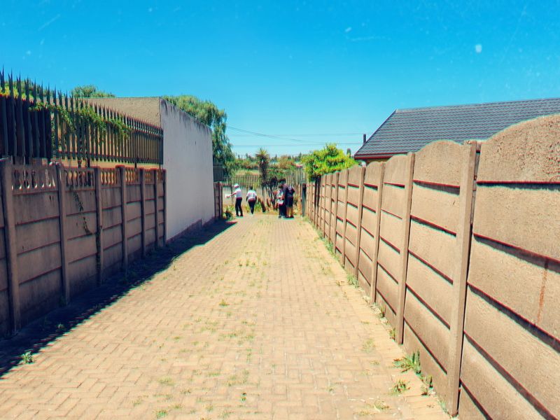 A narrow paved pathway enclosed by precast concrete boundary walls and topped with metal security spikes.