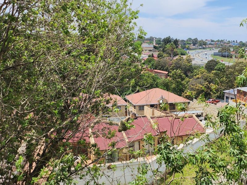 Elevated view overlooking a residential townhouse complex with tiled roofs and surrounding greenery, featuring a distant highway in the background.