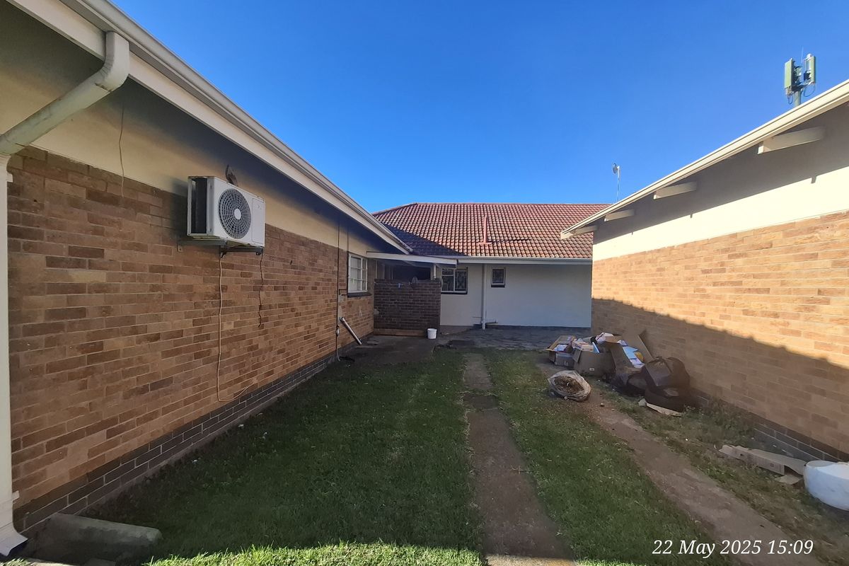 A view of the exterior side passage between two brick buildings, featuring a grass strip, concrete path, and wall-mounted air conditioning unit.