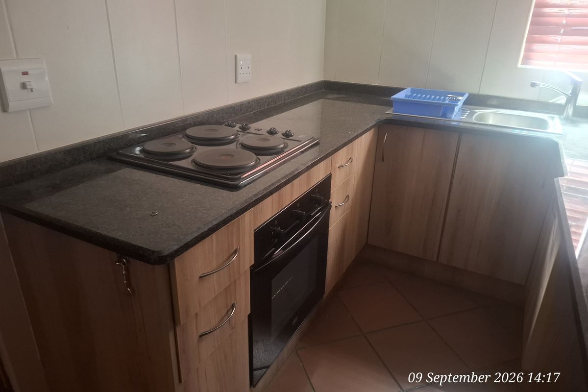 A functional kitchen area featuring wood-finish cabinetry, dark laminate countertops, a built-in electric hob and oven, and a stainless steel sink.