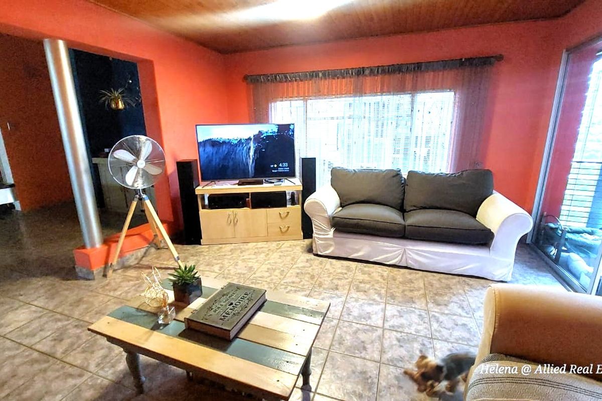 A spacious living room featuring tiled flooring, large windows with security bars, and a timber-paneled ceiling, providing a functional and open living area.