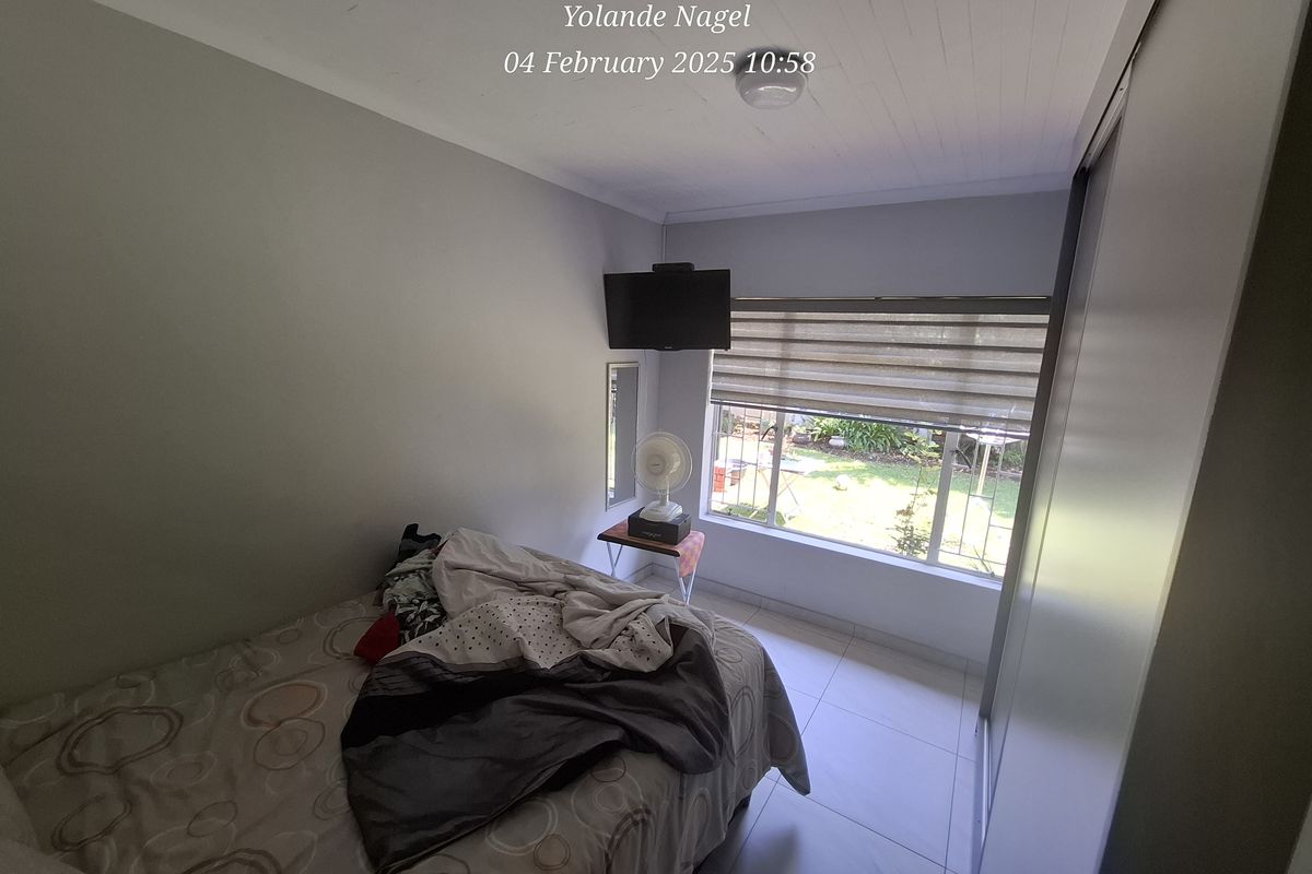 A bedroom featuring tiled flooring, built-in sliding door cupboards, and a window with roller blinds overlooking the garden.