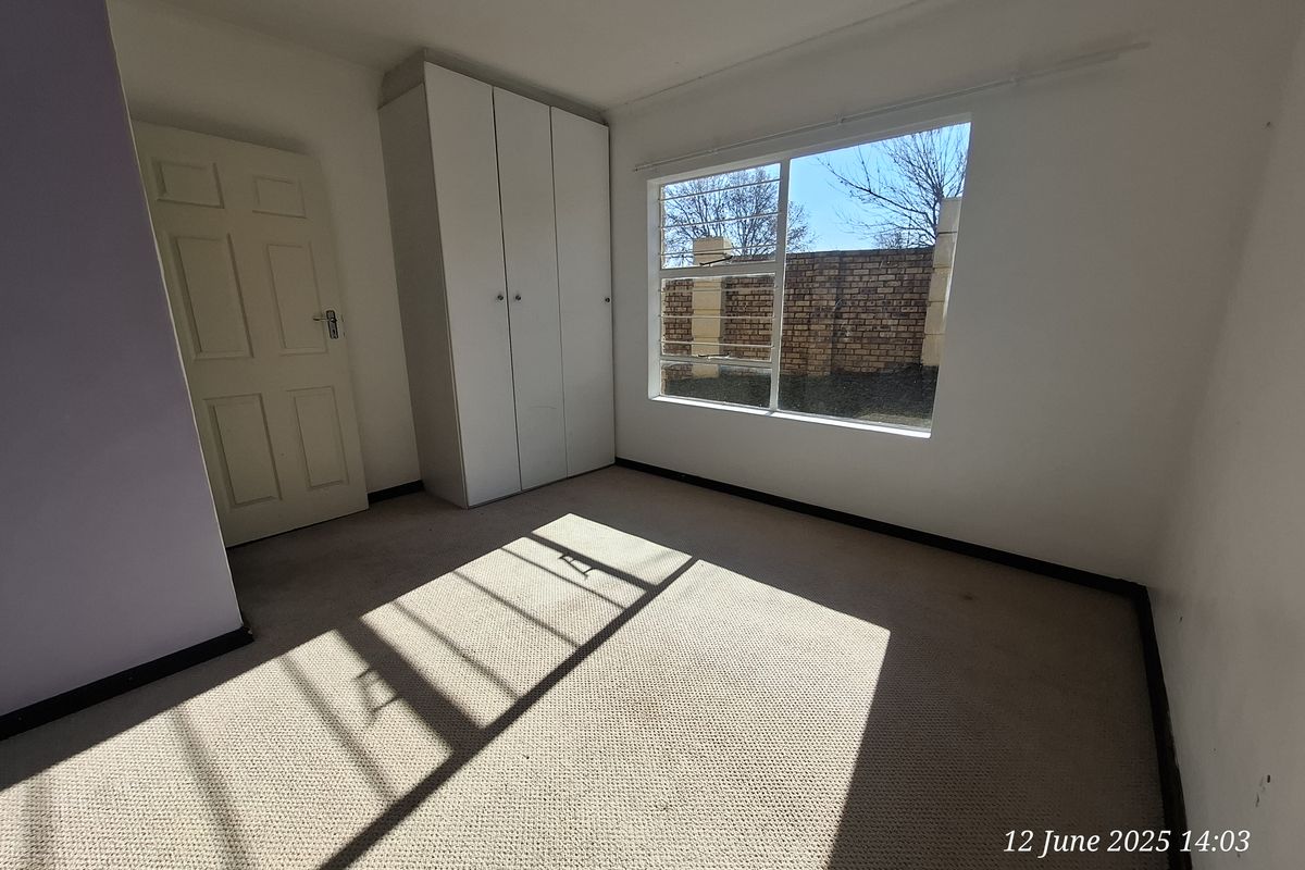 A bright, empty bedroom featuring built-in cupboards, carpeted flooring, and a large window with security bars.