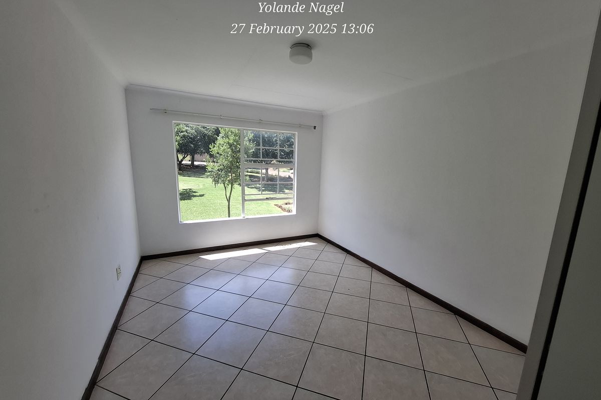A bright, empty bedroom featuring neutral ceramic floor tiles, a large window with garden views, and a simple ceiling-mounted light fixture.