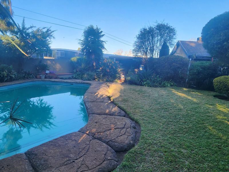 A private backyard featuring a swimming pool with decorative rock-style coping, surrounded by a well-established garden and boundary wall.