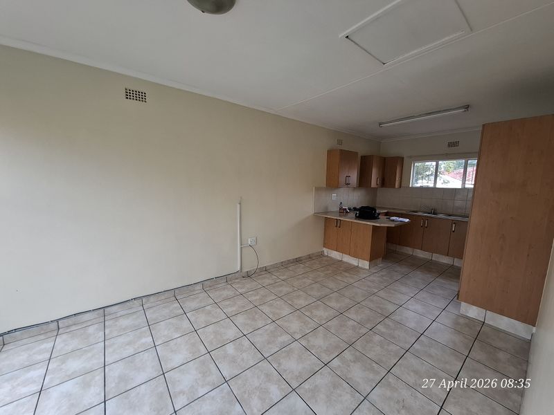 An open-plan living and kitchen area featuring tiled flooring, built-in wooden cabinetry, and a functional layout, presented in a clean, vacant condition.