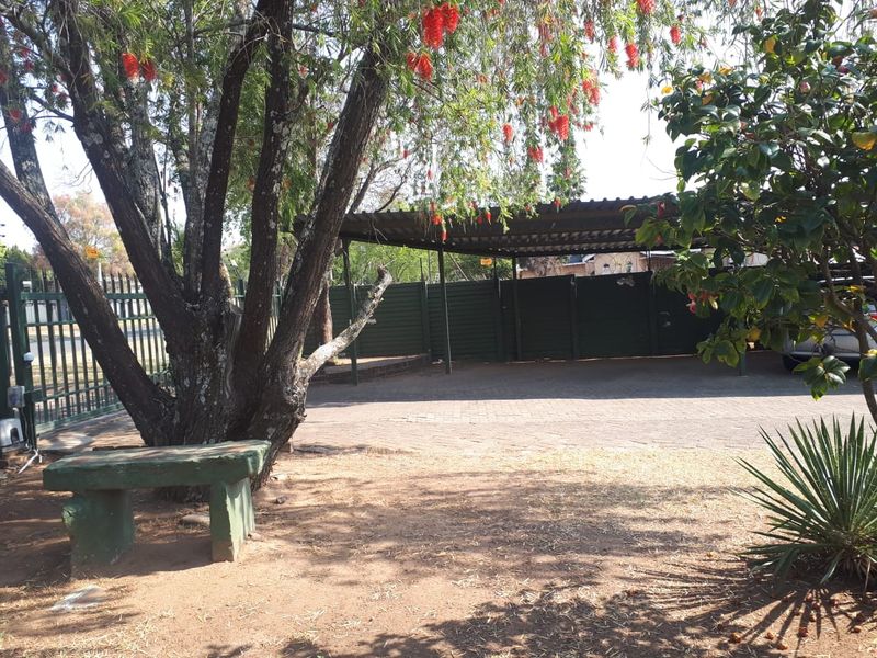 A paved outdoor carport area featuring a metal roof structure, perimeter fencing, and mature trees providing shade.