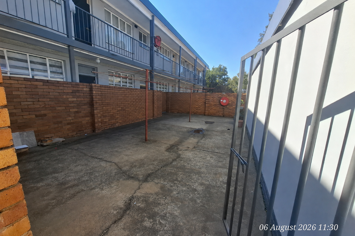 A rear exterior courtyard area featuring concrete paving, brick boundary walls, and a metal security gate, providing functional outdoor space for the flat.