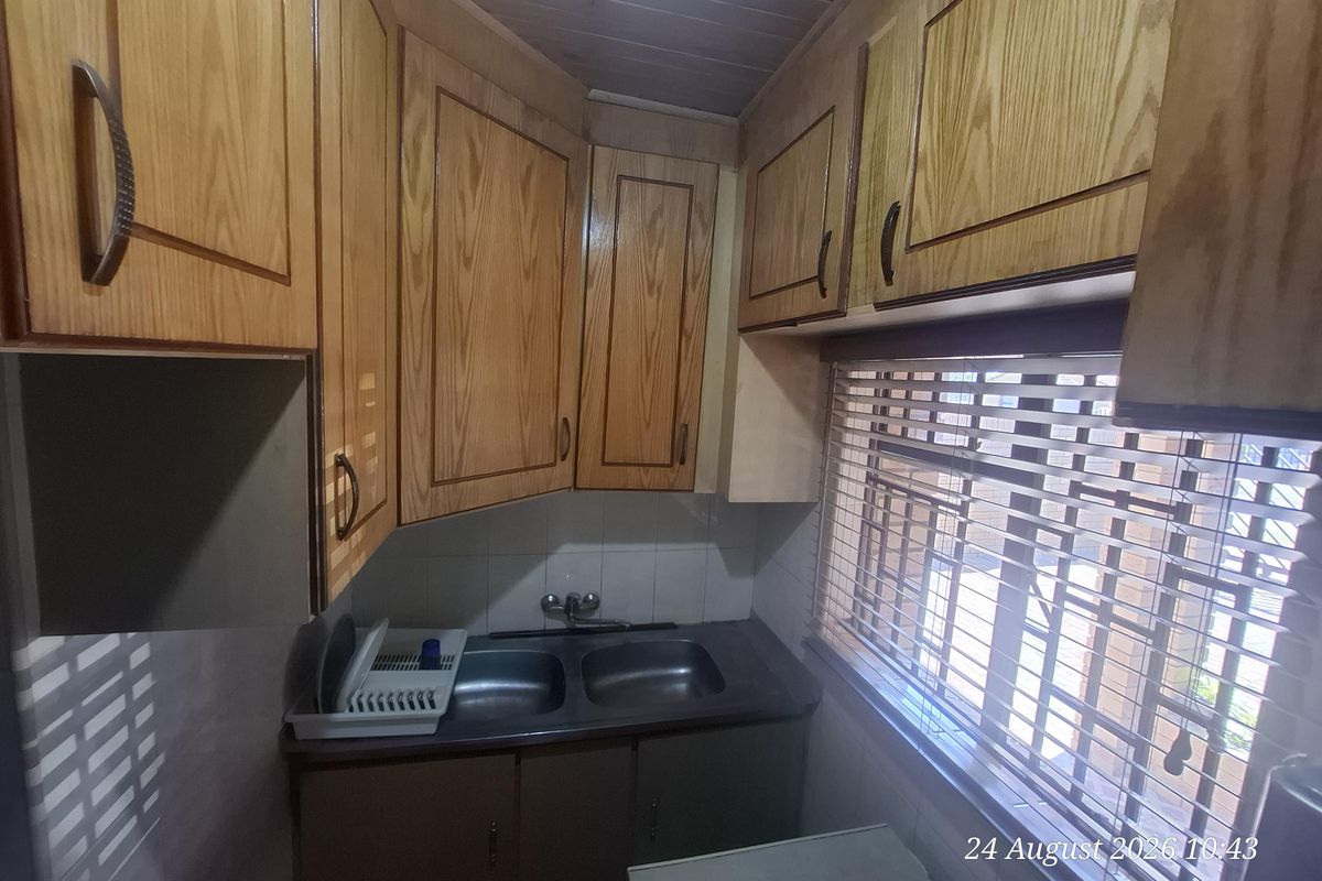A compact kitchen featuring dated wooden cabinetry, a double bowl stainless steel sink, and a window with venetian blinds.
