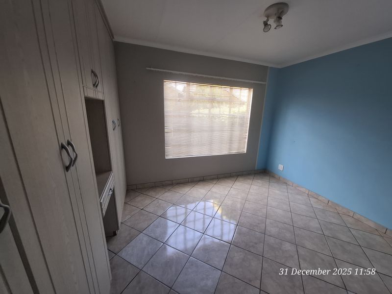 A bright, empty bedroom featuring tiled flooring, built-in cupboards with a vanity section, and venetian blinds on the window.