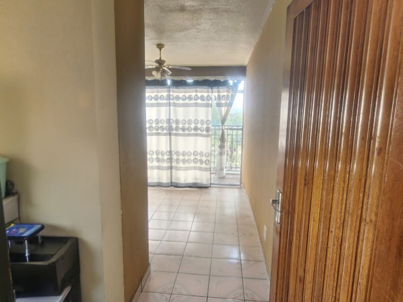A tiled hallway leading towards a balcony with a sliding glass door and ceiling fan, showing dated finishes.