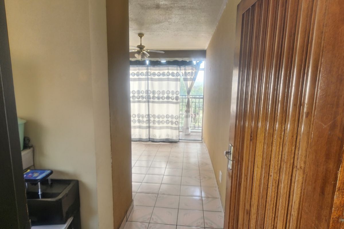 A tiled hallway leading towards a balcony with a sliding glass door and ceiling fan, showing dated finishes.