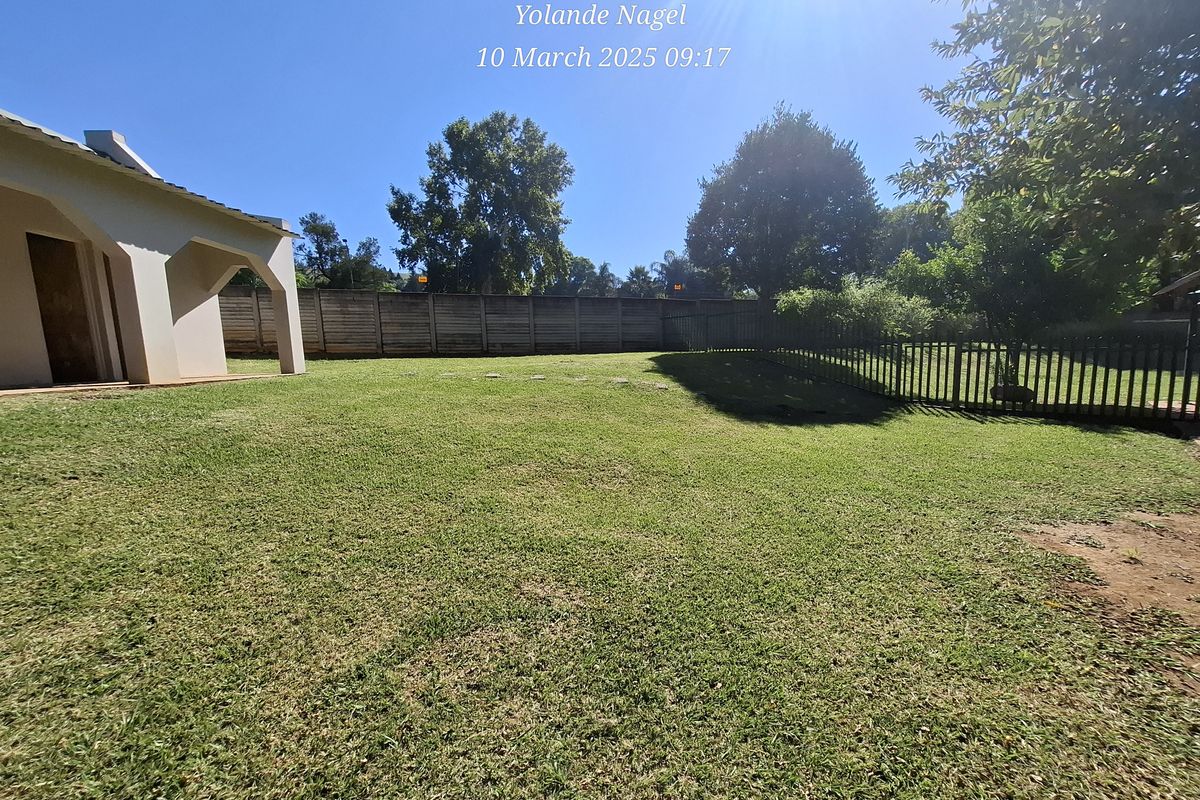 A spacious rear garden featuring a well-maintained lawn, concrete boundary wall, metal fencing, and a covered patio area attached to the main house.