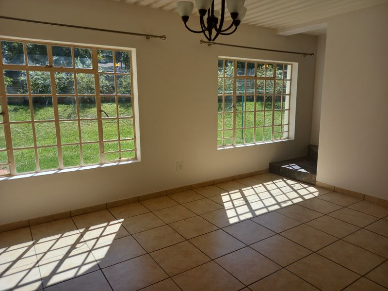 A bright, empty living area featuring neutral tiled flooring and large steel-framed windows overlooking the garden.